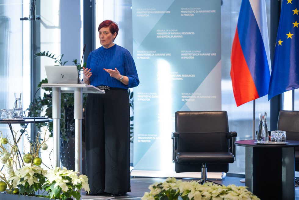 Oseba v modri obleki stoji za govorniškim pultom v dvorani in govori. 