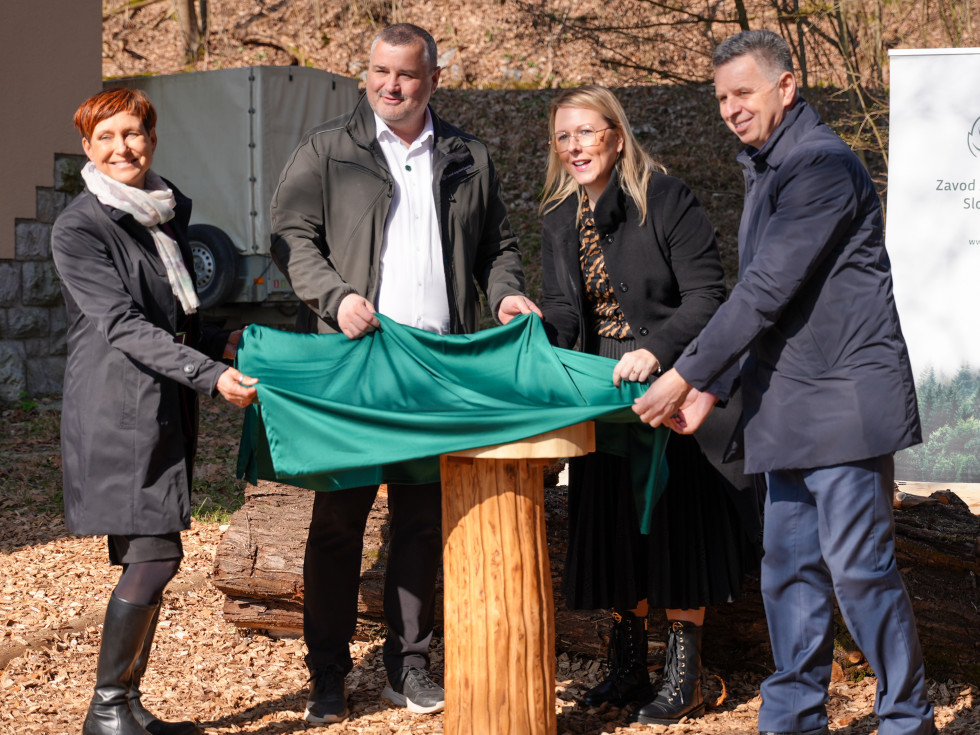 Ministrica skupaj s tremi osebami dviguje prt s koluta pred stavbo