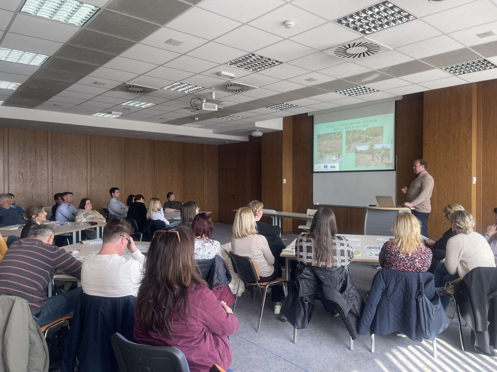 Več oseb sedi v dvorani in posluša predavanje. 