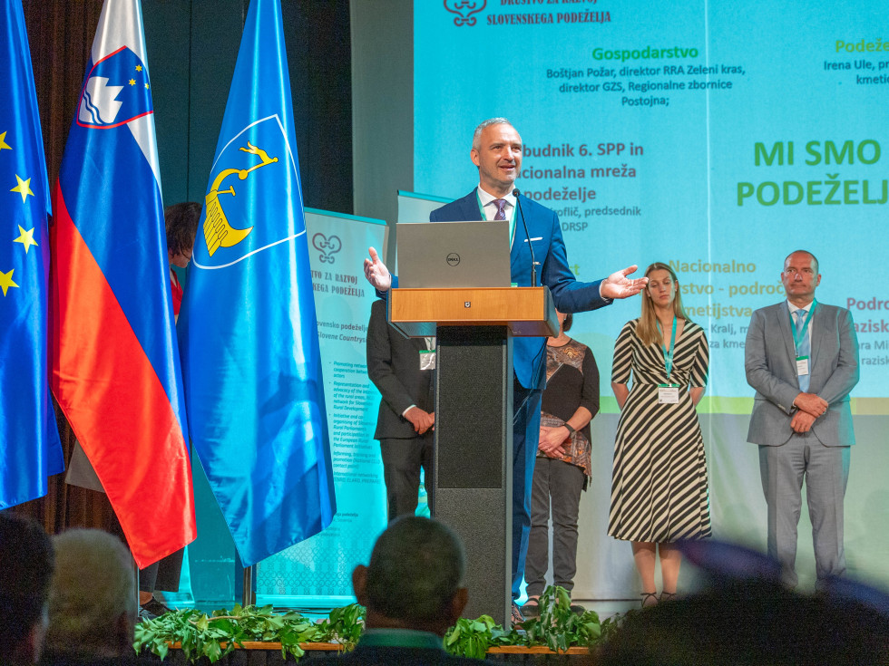 Minister stoji za govorniškim pultom in govori. 