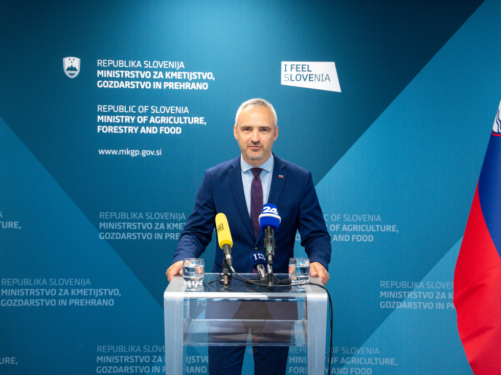 Oseba v modri obleki stoji za govorniškim pultom in govori. 