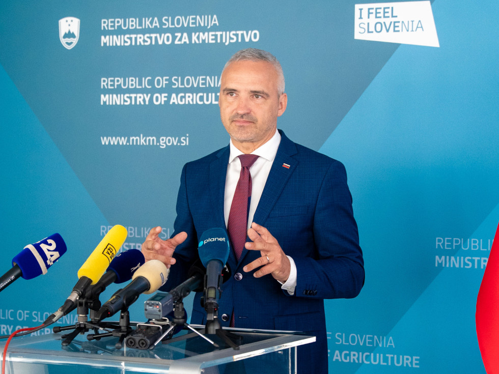 Minister Cigler Kralj stoji za govorniškim pultom, na katerem je več mikrofonov raznih medijev in govori