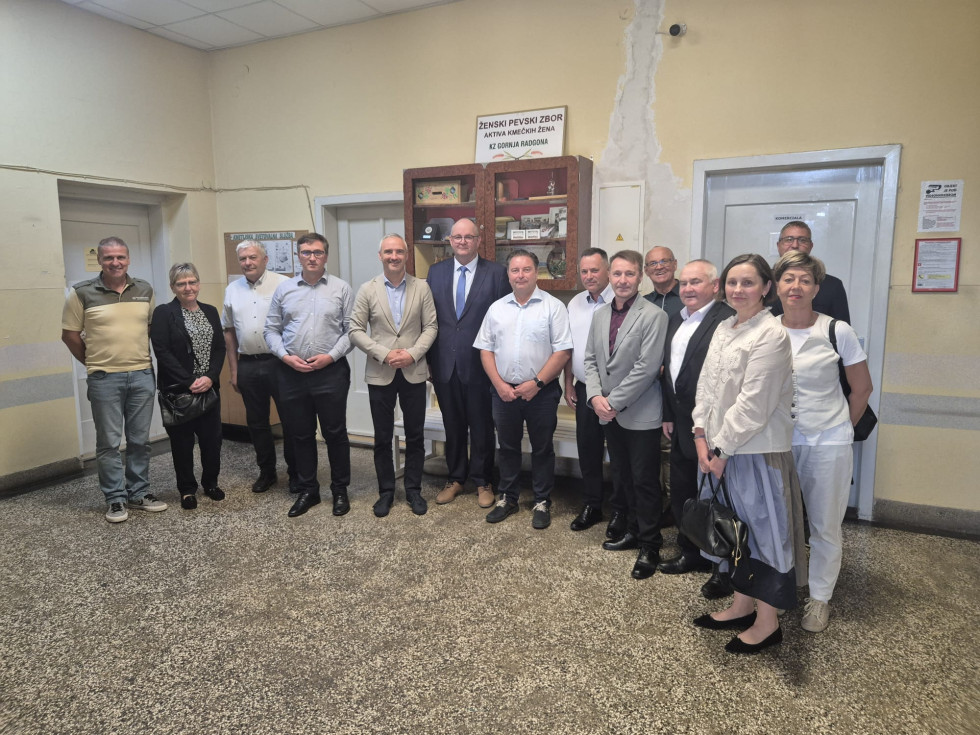 Skupinska fotografija približno petnajstih udeležencev ob obisku ministra v notranjih prostorih stavbe. Udeleženci stojijo v vrsti in gledajo proti fotoaparatu.