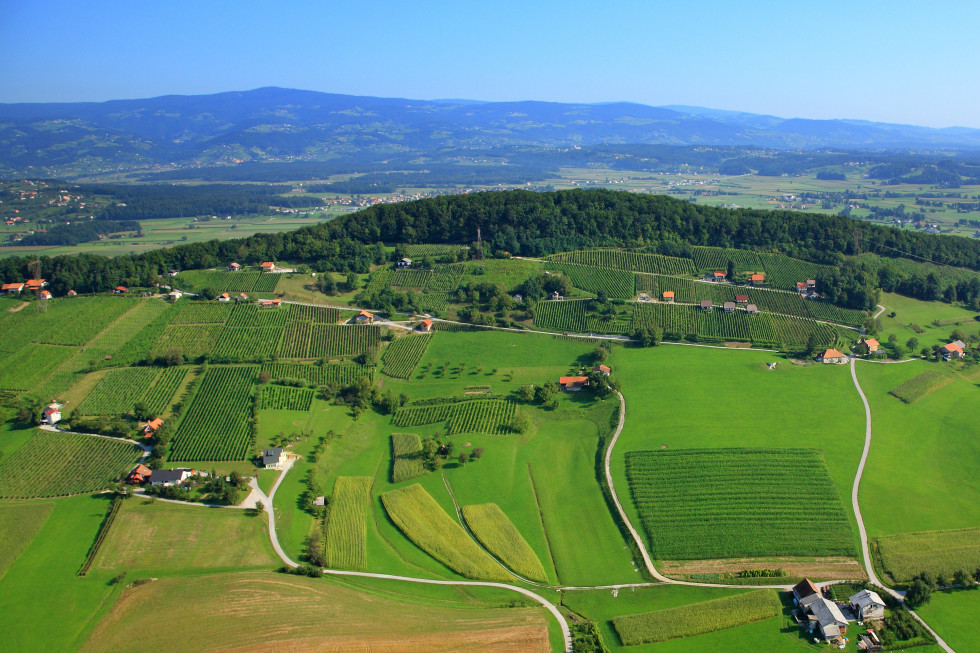 Iz zraka slikana krajina. Vidni so zeleni travniki in gruče hiš ter v daljavi hribovje.
