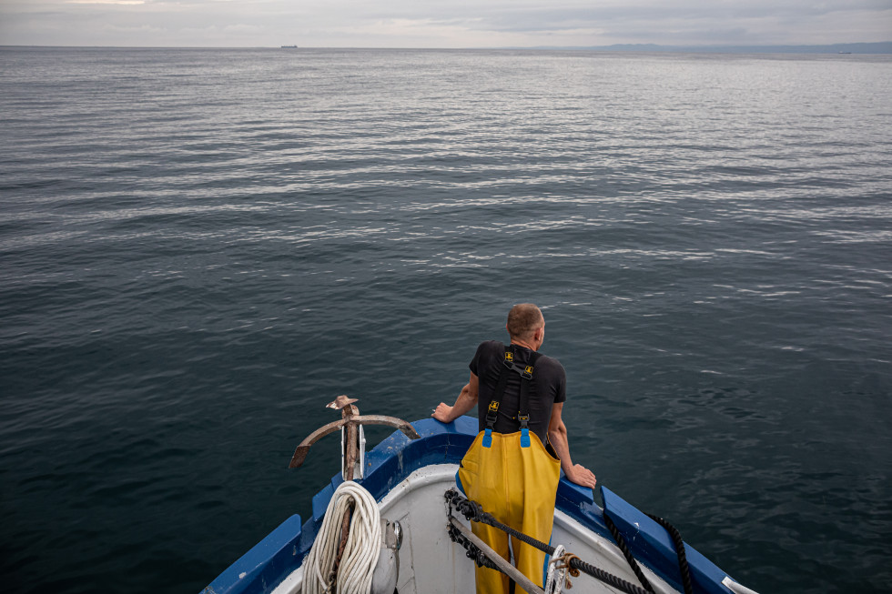 Konica ribiške ladje na kateri stoji ribič v rumenih hlačah in zre v širno morje.