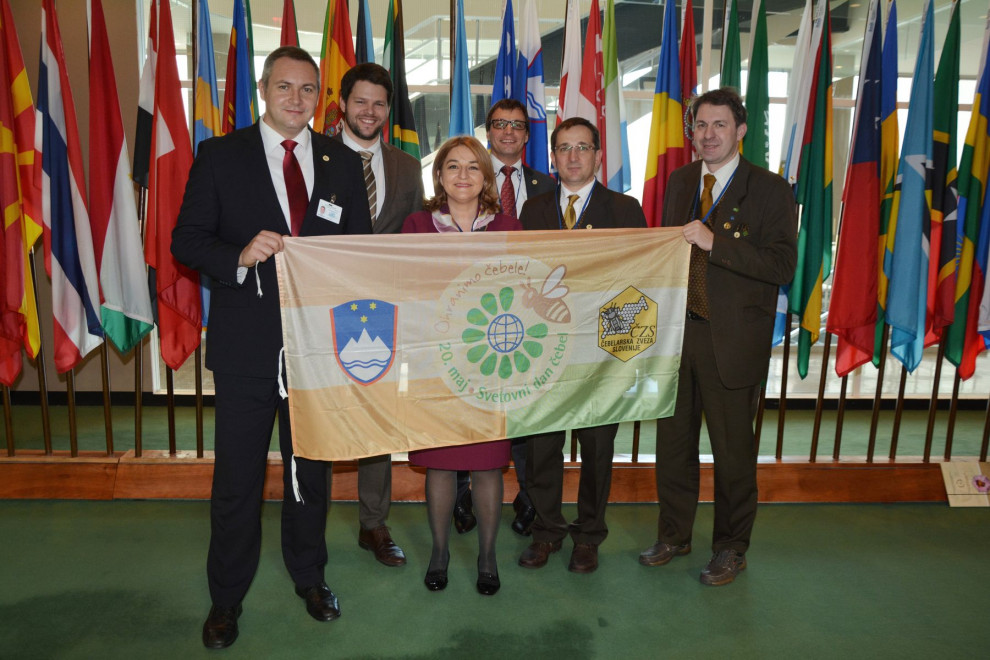 Skupina ljudi v rokah drži zastavo s slovenskim grbom in logotipom svetovnega dne čebel