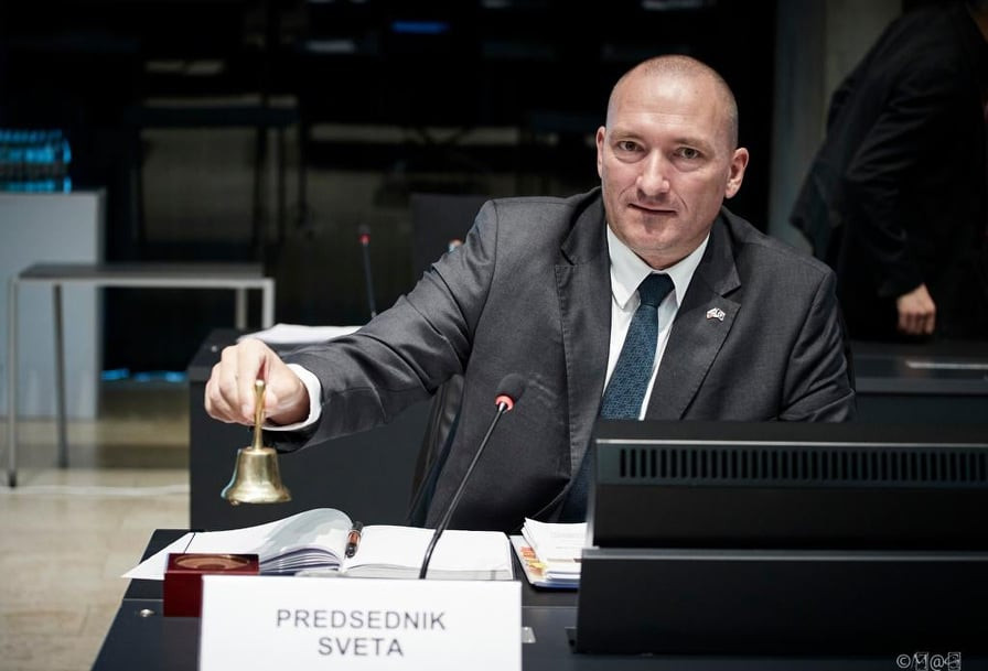 Minister ob začetku sveta. V rokah ima zvonček s katerim pozvoni za začetek zasedanja. 