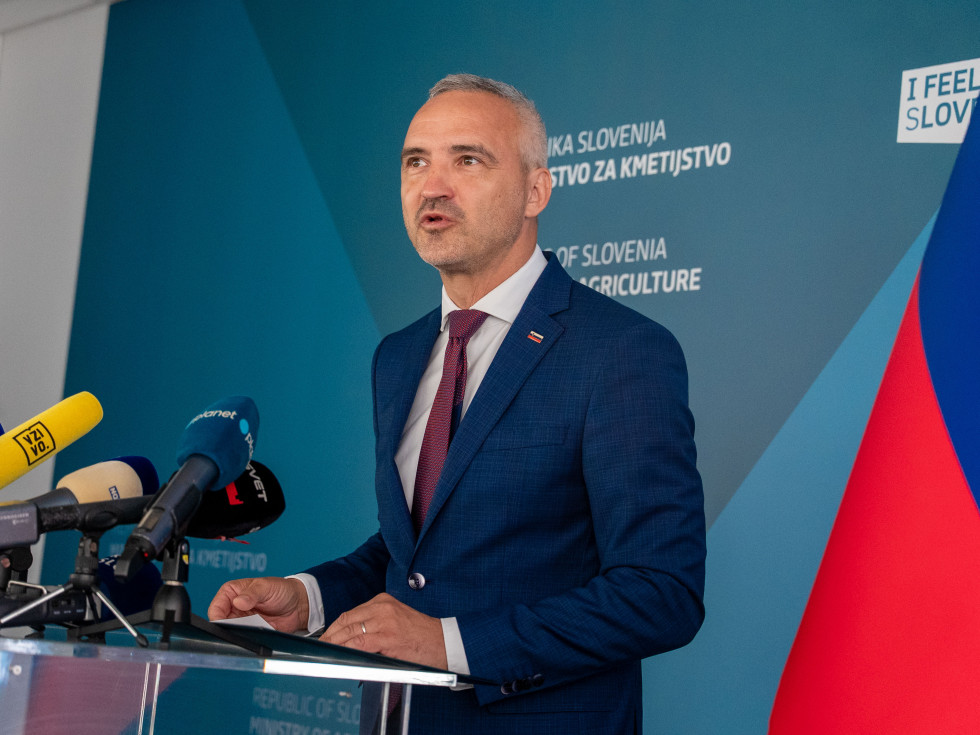 Minister oblečen v modro obleko stoji za govorniškim pultom in govori. 
