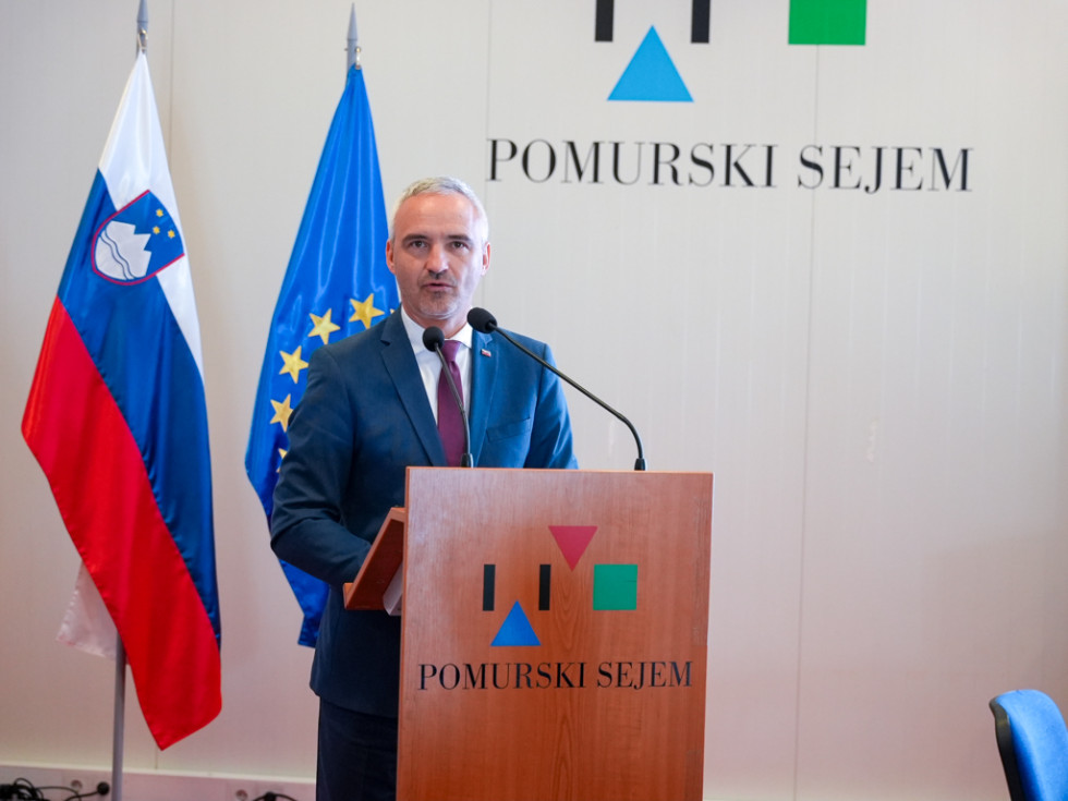 Oseba stoji za govorniškim pultom in govori. 