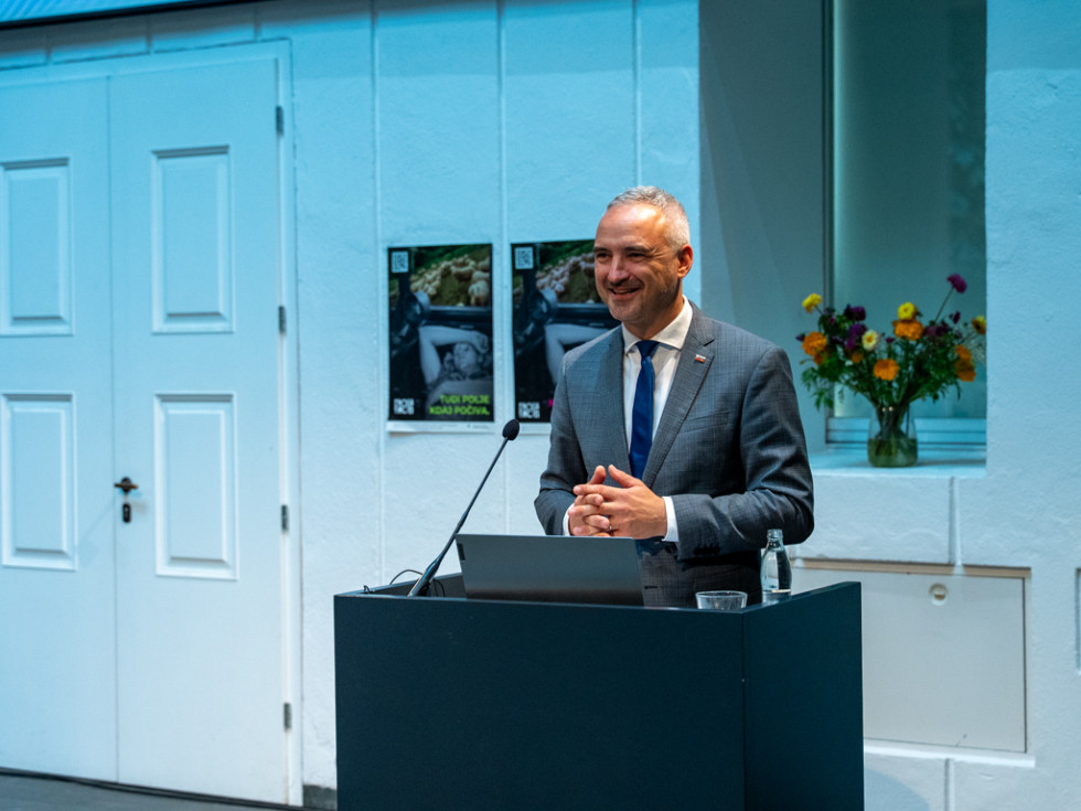 Minister stoji za govorniškim pultom in govori. 