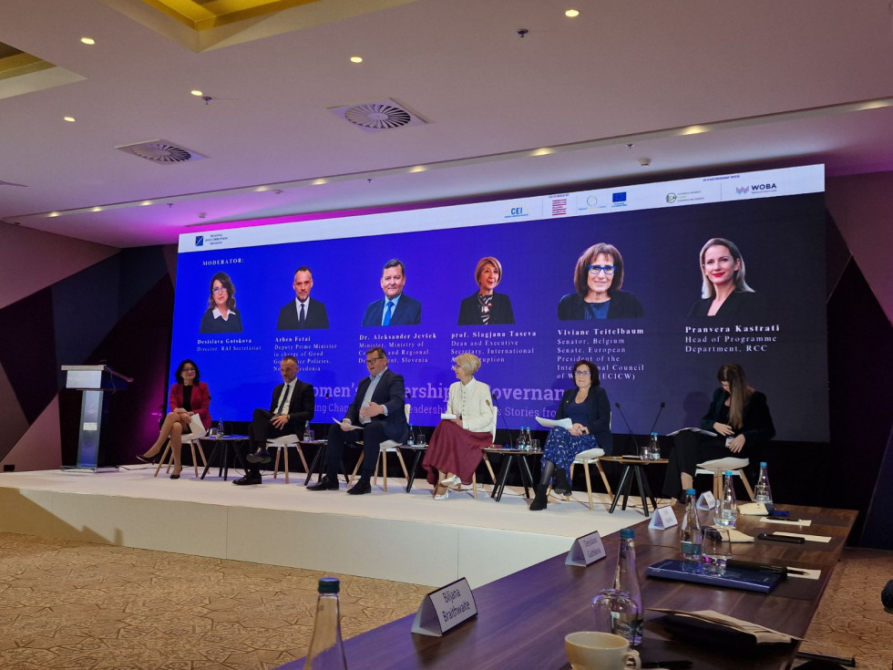 Four women and two men are seated on chairs facing the audience, with their names and functions and photographs on a screen behind them.