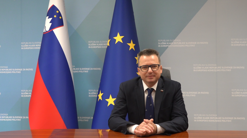 Moški sedi pred mizo s sklenjenim rokami, na njegovi desni strani sta slovenska in evropska zastava.