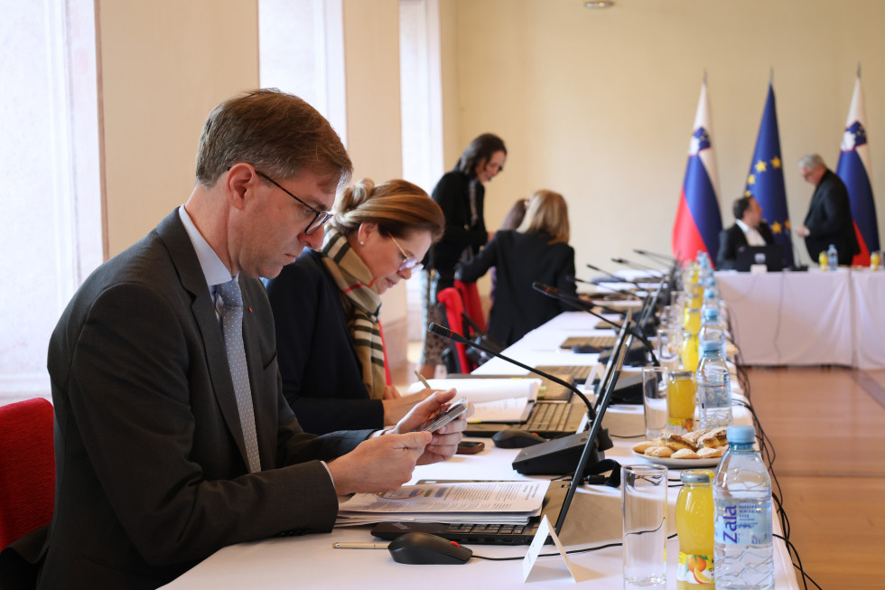  Moški in ženske sedijo za dolgo mizo. Na mizi mikrofoni in prenosni računalniki.