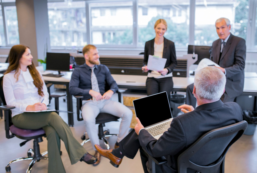 In the foreground, an older man faces his team at a meeting.