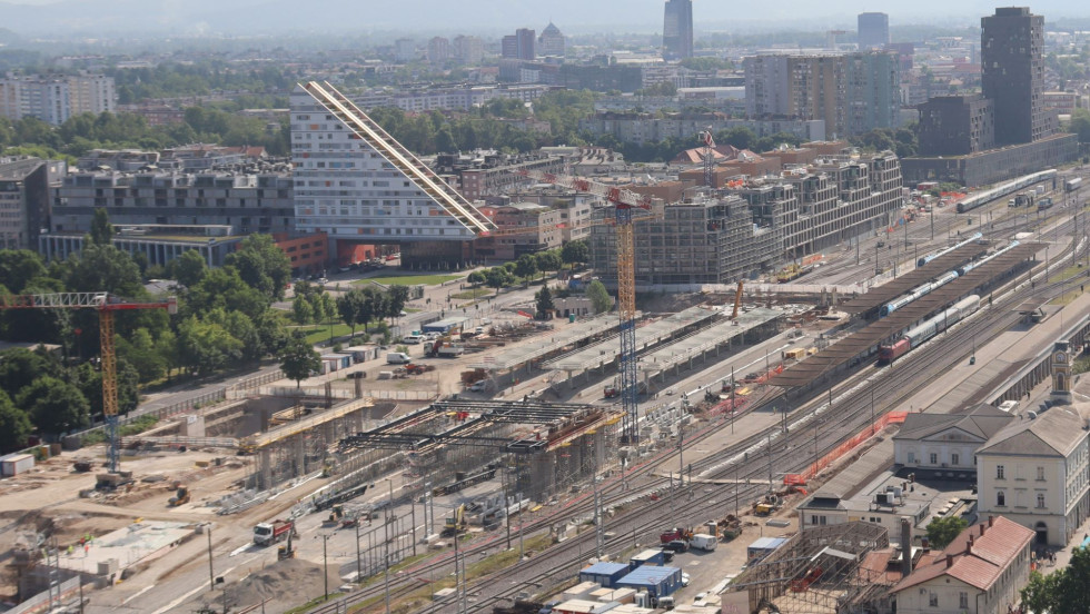 A bird's-eye view of the construction site of the Ljubljana railway station. In the foreground, you can see the tracks, a construction crane, and work on the new overpass. Ljubljana is in the background.
