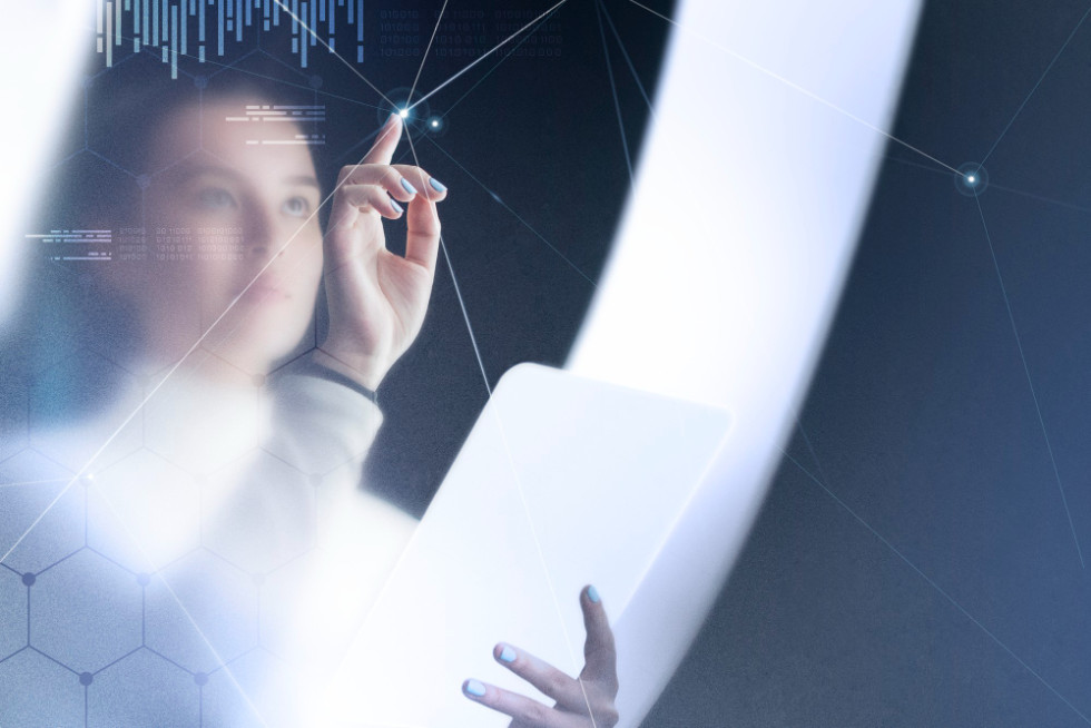 Ženska stoji pred lebdečim, prosojnim virtualnim zaslonom, na katerem so prikazane svetleče digitalne ikone, povezave in podatkovni tokovi. Z rokami se dotika navideznih elementov, kot da upravlja napredno tehnologijo v sodobnem, visokotehnološkem okolju.