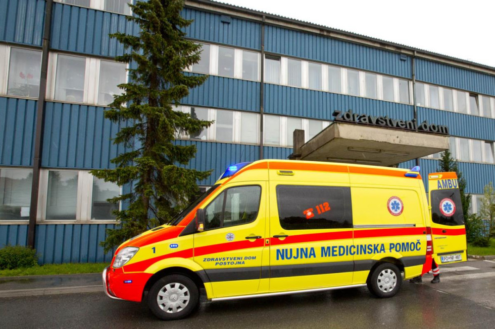 V ospredju reševalno vozilo, parkirano pred zdravstvenim domo, Stavba zdravstvenega doma vidna v ozadju. 