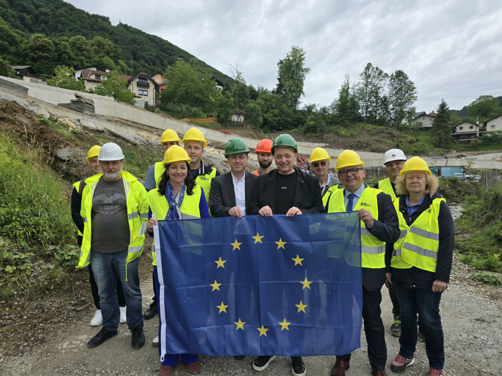 Skupina udeležencev v zaščitni opremi drži zastavo Evropske unije na terenu gradbenega oziroma infrastrukturnega projekta.