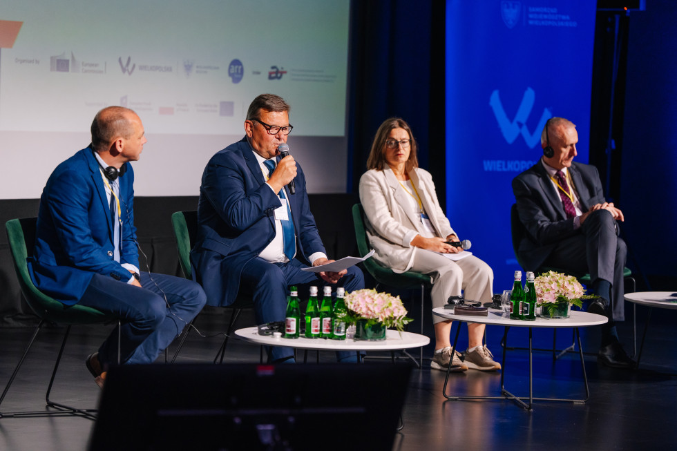 Trije moški in ženska sedijo v vrsti na odru, pred njimi majhne mizice. Drugi z leve strani minister Jevšek im a v roki mikrofon in govori. 