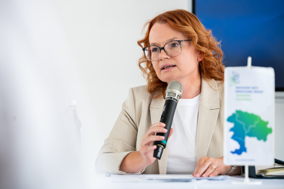 Gospa sedi za mizo in govori na mikrofon.