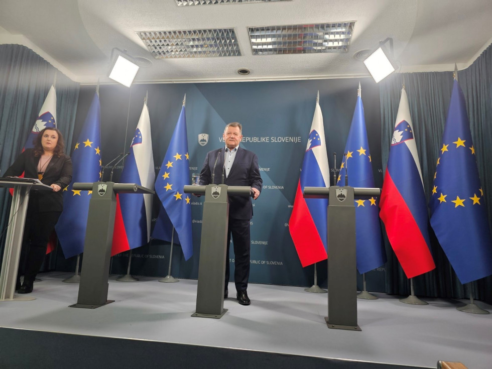 Minister Jevšek na novinarski konferenci v tiskovnem središču vlade stoji za govorniškim pultom. Zadaj slovenske in evropske zastave.