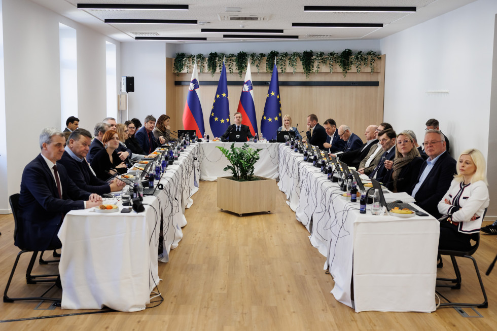 Ministri sedijo za omizjem pred pričetkom seje vlade. Na mizah prenosni računalniki in mikrofoni. Na koncu prostora slovenski in evropski zastavi. 