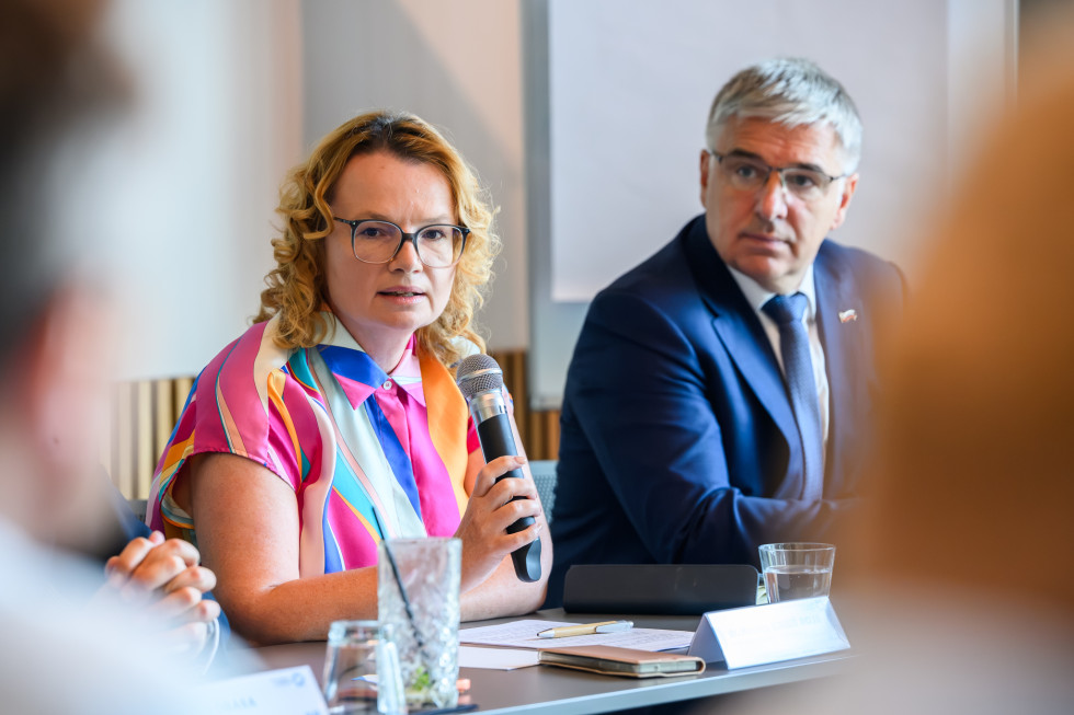 Ženska za omizjem govori, v ozadju za istim omizjem udeleženec.