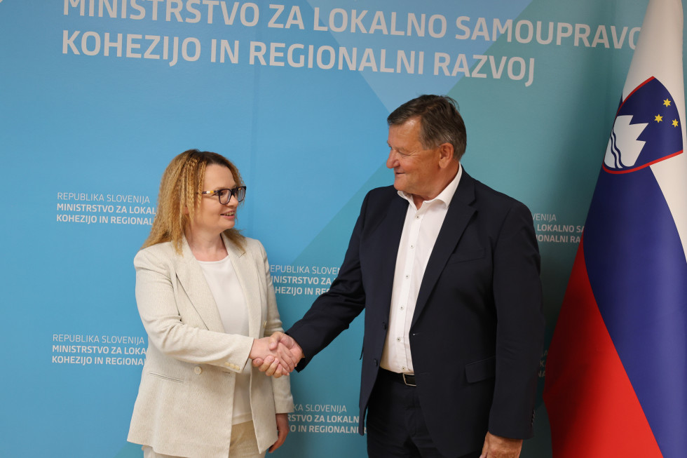 A woman and a man shake hands in front of a sign reading "Ministry of Cohesion and Regional Development."
