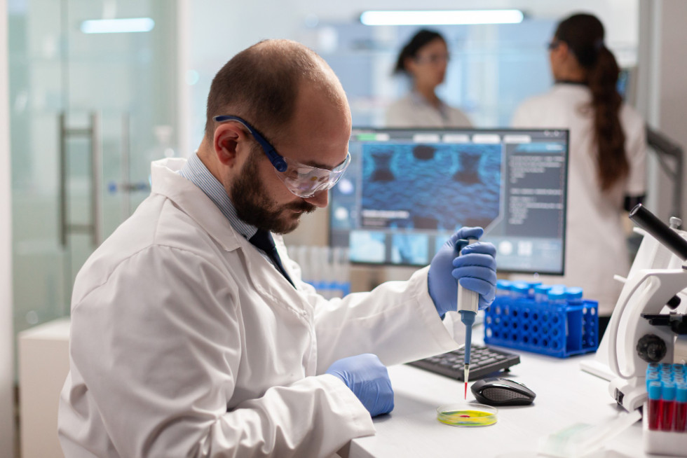 Biokemik v laboratoriju analizira vzorec krvi. Oblečen je v zaščitno haljo in rokavice ter z mikropipeto natančno odmerja majhno količino tekočine v laboratorijsko posodico.
