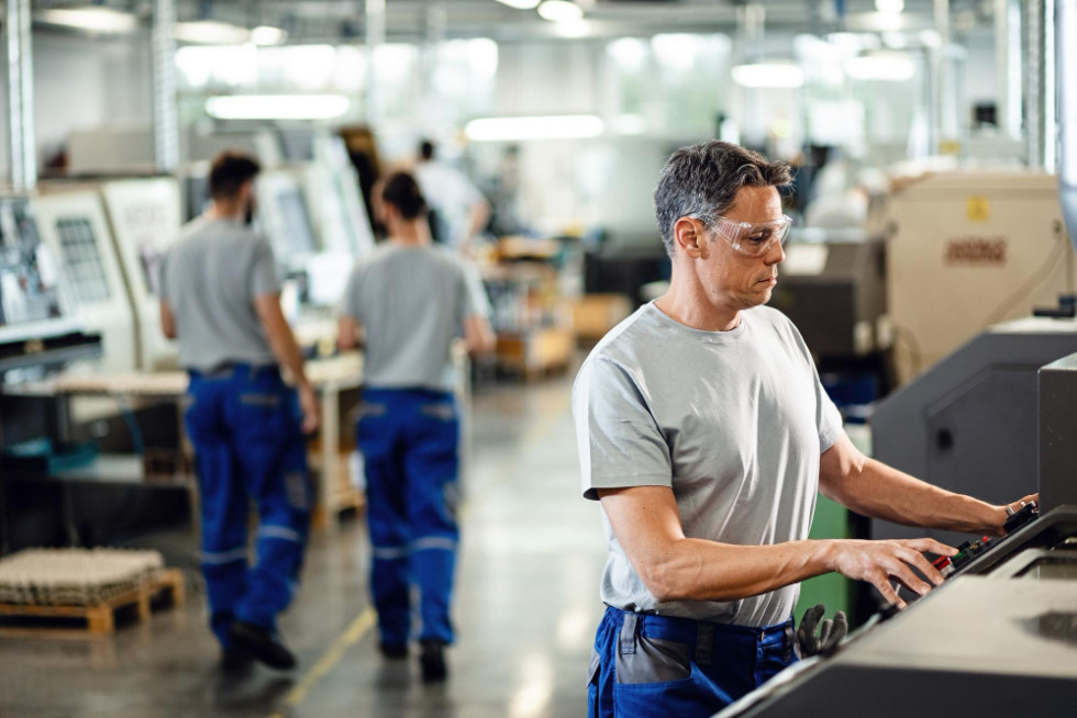 Delavec v industrijskem obratu upravlja CNC stroj. Stoji ob napravi in nadzoruje proces, okoli njega je vidna industrijska oprema in proizvodno okolje.