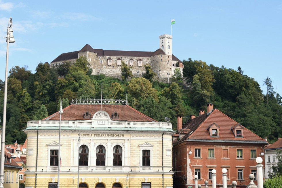 Pogled na Ljubljanski grad in grajski grič z drevesi, pod njim oranžna in rumena stavba (Slovenska filharmonija)