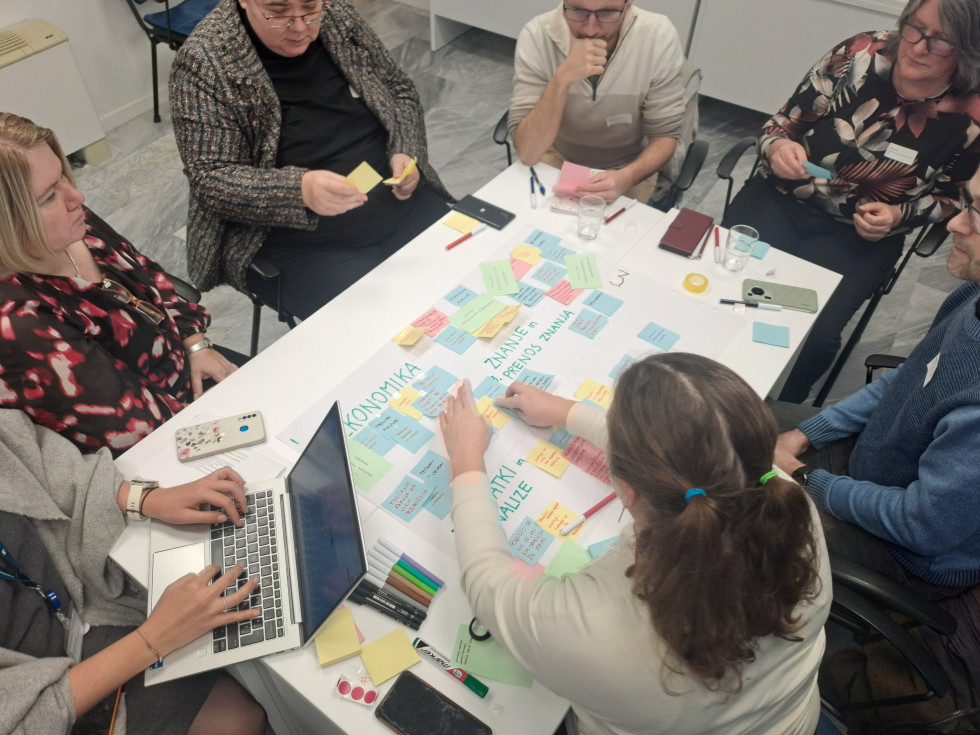 Udeleženci delavnice sedijo okoli mize in na sredo lepijo ideje na stickerjih