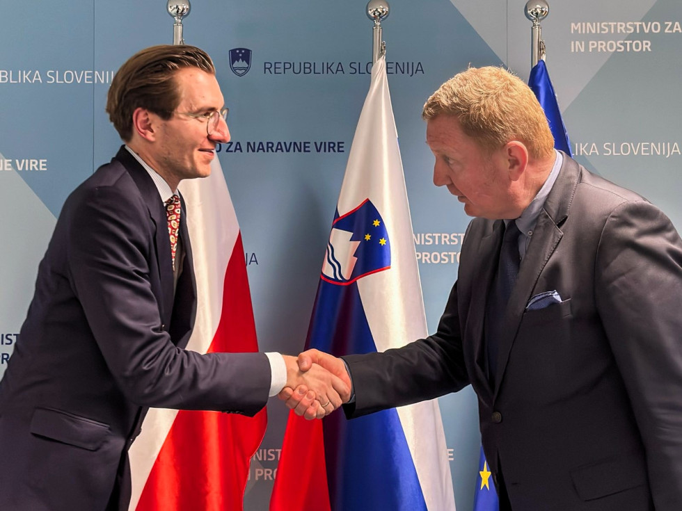 Državna sekretarja Szyska in Gajšek stojita pred poljsko, slovensko in evropsko zastavo ter se rokujeta