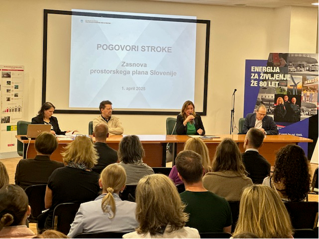 V veliki sejni sobi udeleženci poslušajo govorce, ki sedijo spredaj za mizo in predstavljajo zasnovo prostorskega plana Slovenije