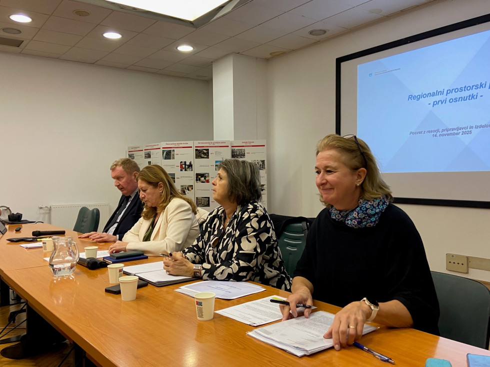 Moški in tri ženske sedijo za govorniško mizo. Pred sabo imajo papirje. Zadaj predstavitev.