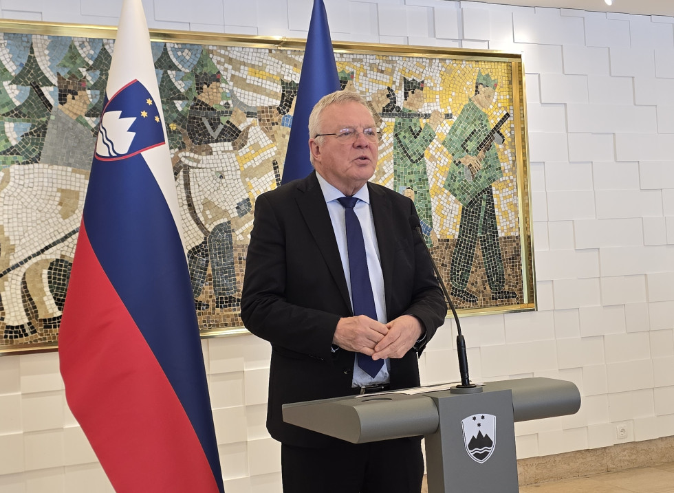 Minister Novak za govornico na novinarski konferenci, ob njem zastave, za njim stenski mozaik s podobo partizanskega boja