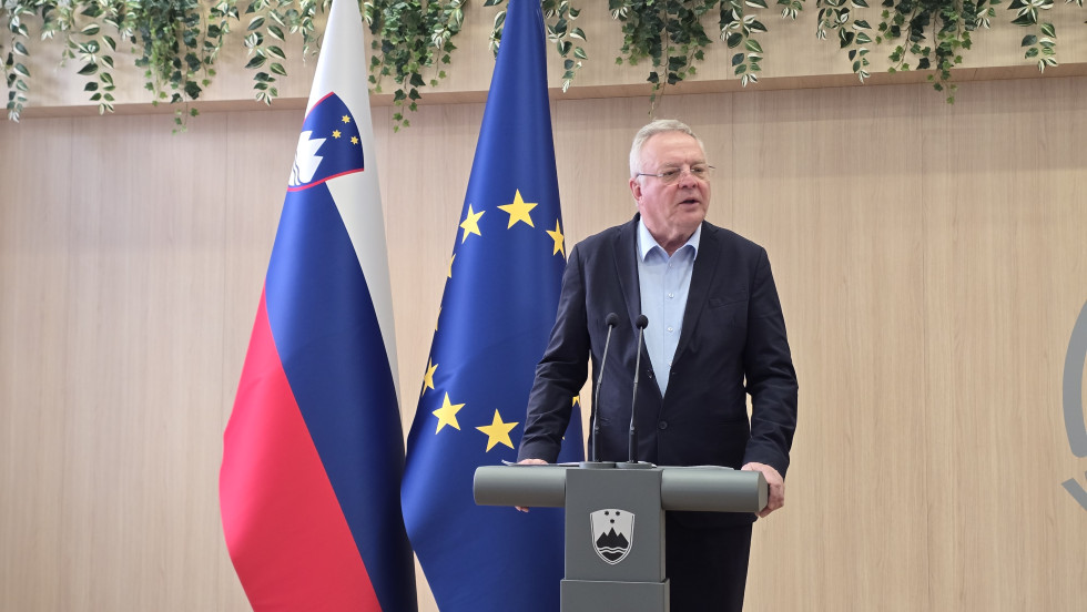 Minister za govornico nagovarja novinarje. Za njim slovenska in evropska zastava.