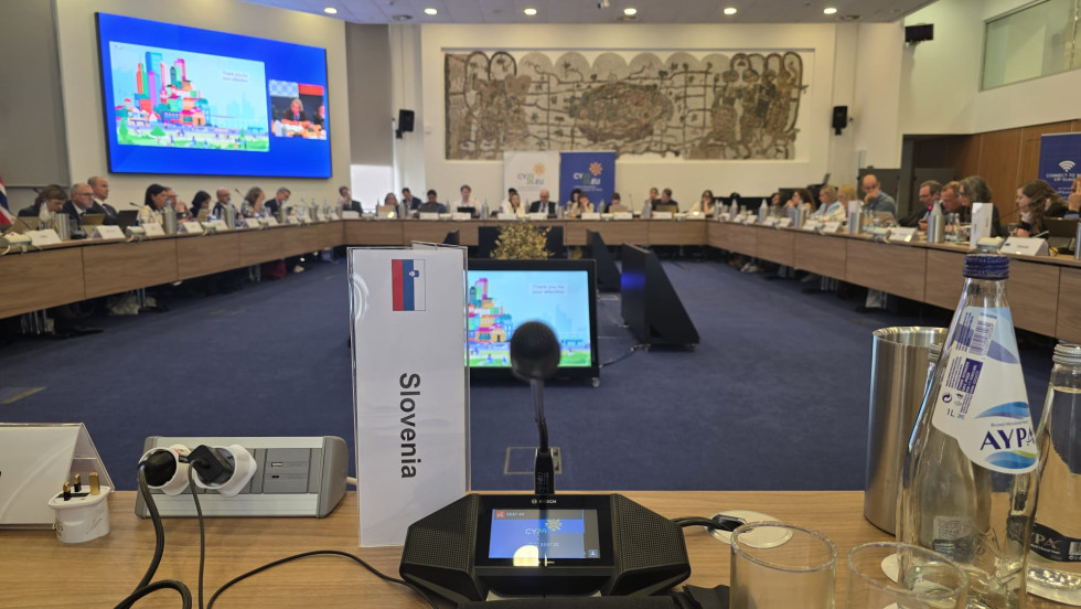 Dvorana, mize, udeleženci sedijo, pred njimi tablice z imeni in mikrofoni.