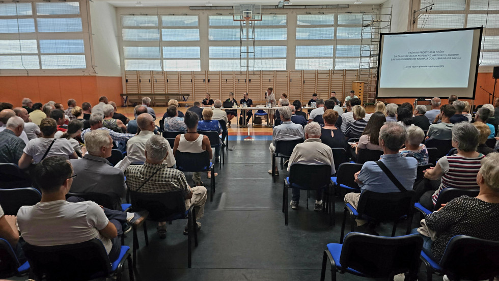 Udeleženci javne predstavitve sedijo v dvorani in spremljajo govorce na odru