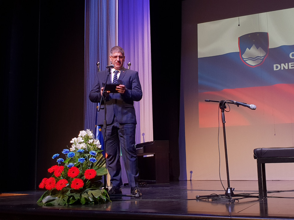 Minister za notranje zadeve Boštjan Poklukar pri nagovoru stoji na odru, v ozadju na zaslonu slovenska zastava.