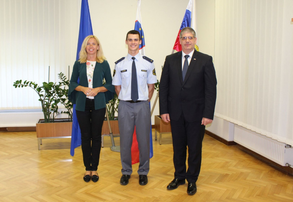 Generalna direktorica policije mag. Tatjana Bobnar, Tim Gajser v policijski uniformi in notranji minister Boštjan Poklukar stojijo pred zastavami.