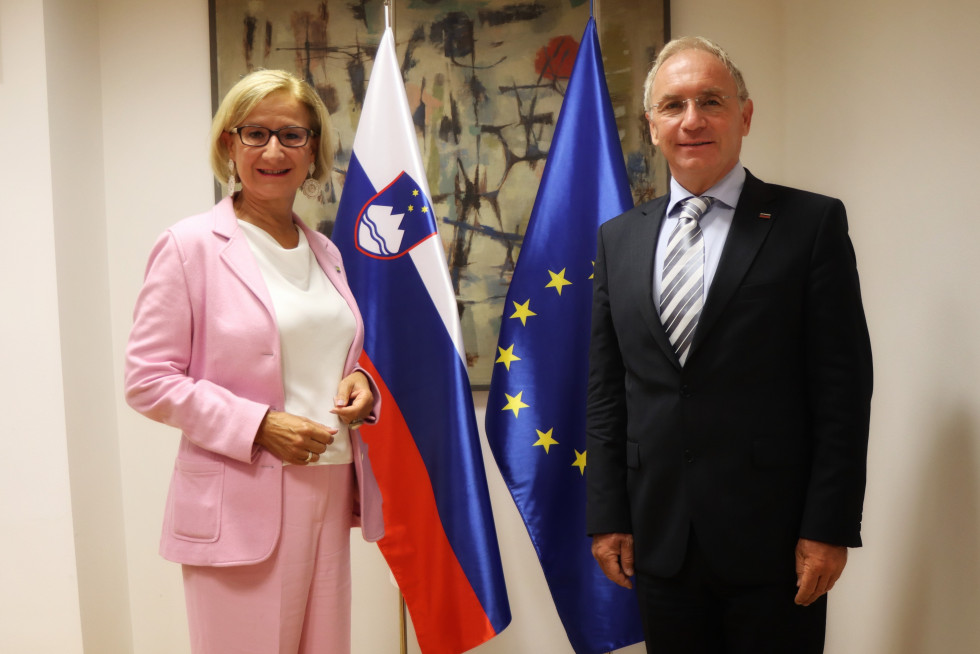 Minister Aleš Hojs in deželna glavarka Spodnje Avstrije Johanna Mikl-Leitner stojita pred zastavo EU in zastavo Slovenije, na steni je umetniška slika.