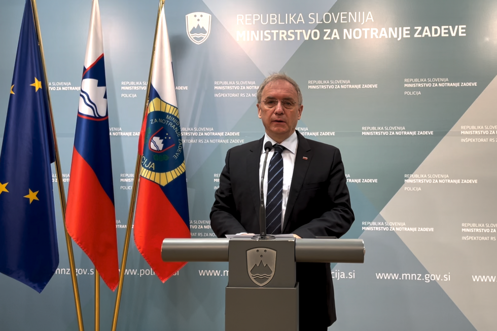 Minister za notranje zadeve Aleš Hojs stoji za govornico, pred modrim ozadjem z napisi Ministrstvo za notranje zadeve in zastavami.