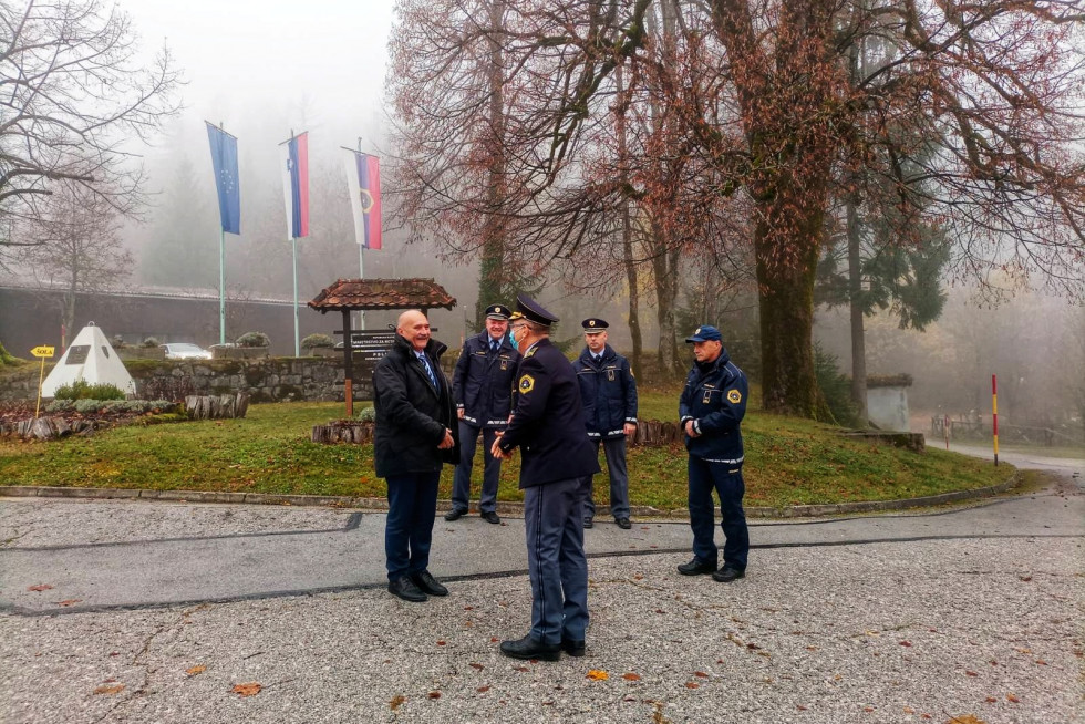 Vadbeni center Gotenica, policisti v uniformah in državni sekretar stojijo v krogu, zadaj zastave, megleno.