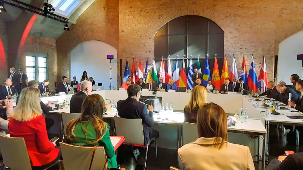 The meeting participants are seated at a table set up in a U-shape, with flags and a brown background behind them