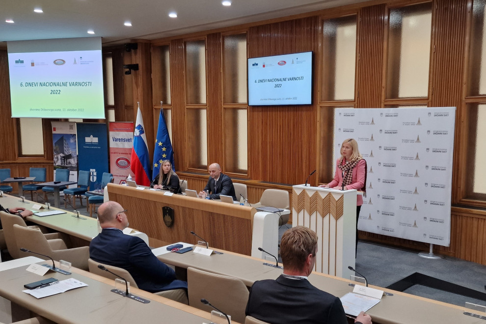 Ministrica Tatjana Bobnar stoji za govorniškim pultom, za njo ozadje belo-rjave barve in ekran z napisom 6. Dan nacionalne varnosti, pred njo udeleženci sedijo za mizami