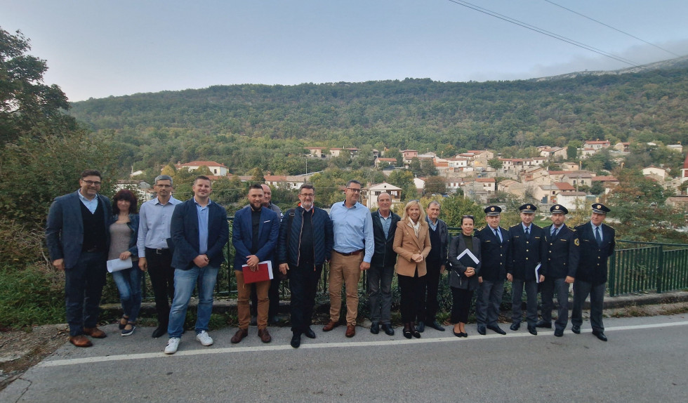 Udeleženci sestanka stojijo zunaj na asfaltu, za njimi gozd, in se slikajo skupinsko