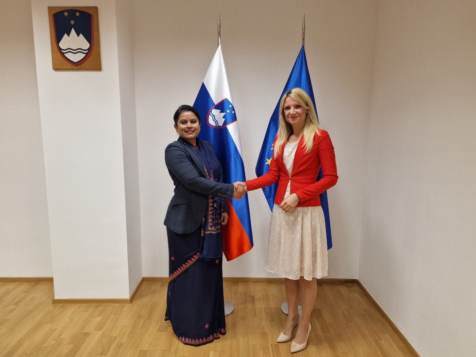 Državna sekretarka Tina Heferle in veleposlanica Indije Namrata Satdeve Kumar stojita pred zastavama Slovenije in EU in se rokujeta