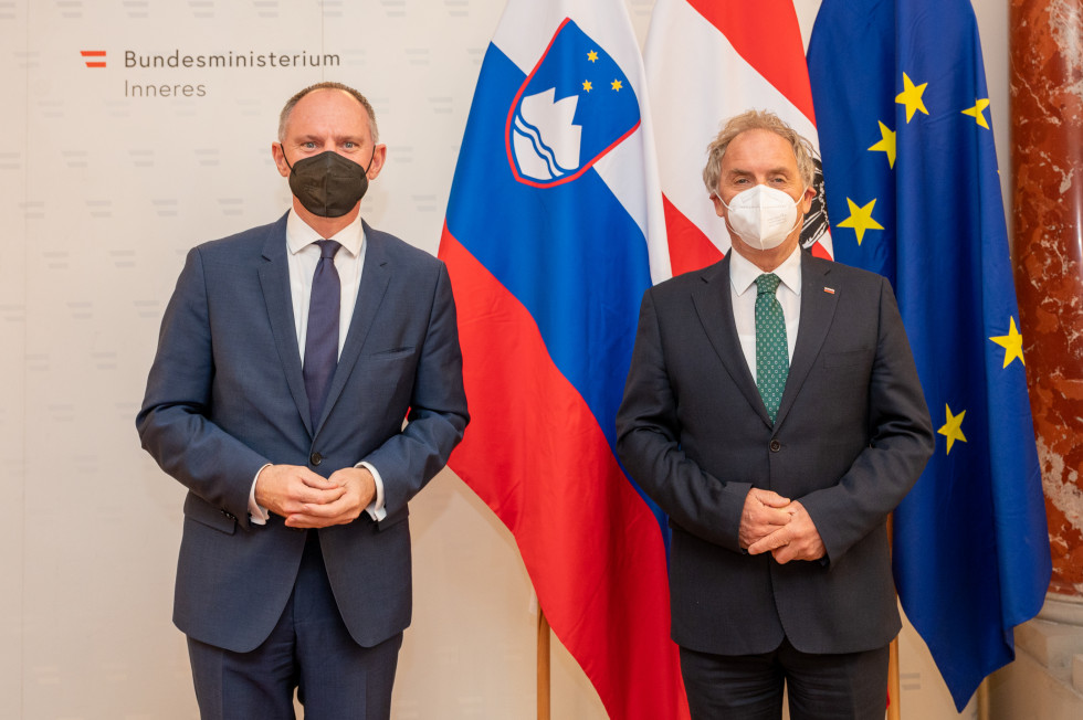 Dvostransko srečanje slovenskega in avstrijskega ministra za notranje zadeve ob robu konference na Dunaju. Ministra Karner in Hojs stojita pred slovensko in avstrijsko zastavo.