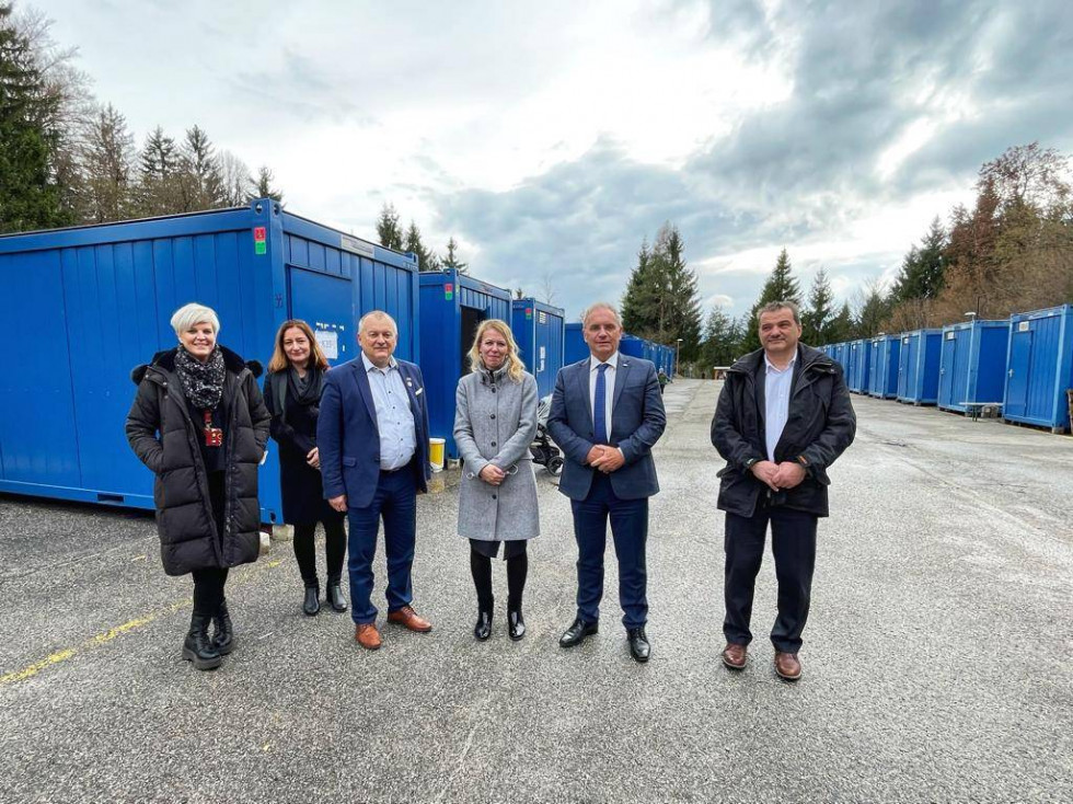 Minister Aleš Hojs in državni sekretar Franc Kangler sta obiskala nastanitveni center v Logatcu, stojijo pred modrimi zabojniki.